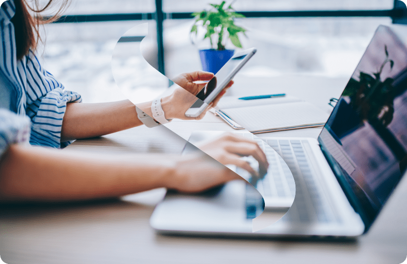 Une personne, un expert-comptable partiellement cachée, est assise à un bureau, une main tapant sur un ordinateur portable et l'autre tenant un smartphone. Un bloc-notes et une plante en pot sont posés sur le bureau. L'arrière-plan présente de grandes fenêtres avec une vue floue de La Ciotat.